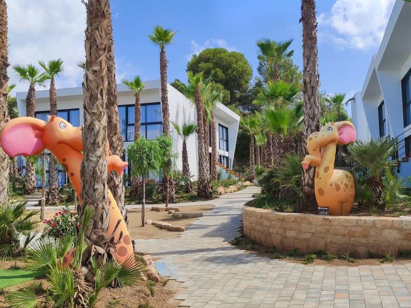 Kostkowana alejka z rzeźbami drewnianych zwierząt i palmami między nowoczesnymi budynkami.