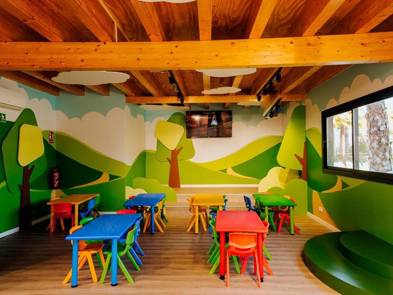 Kolorowa sala zabaw z małymi drewnianymi stołami i krzesłami pod drewnianym sufitem.