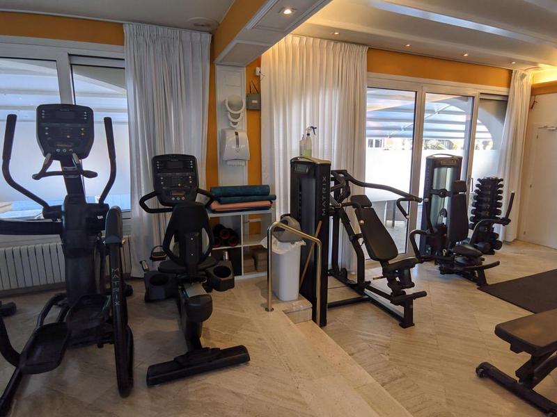 Fitness room with various exercise machines in front of large windows and wooden floor.