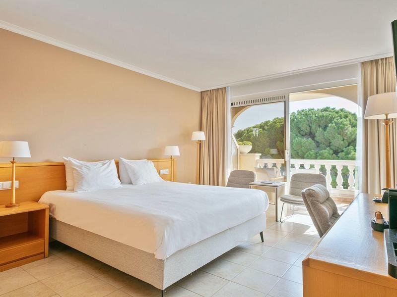 Bright hotel room with double bed, balcony, and view of green trees.