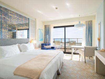 Chambre d'hôtel lumineuse avec grand lit, bureau et vue sur la ville par porte coulissante en verre.