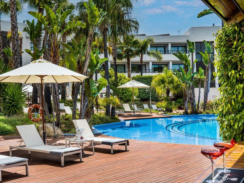 Espace piscine de l'hôtel avec parasols, transats et plantes tropicales.
