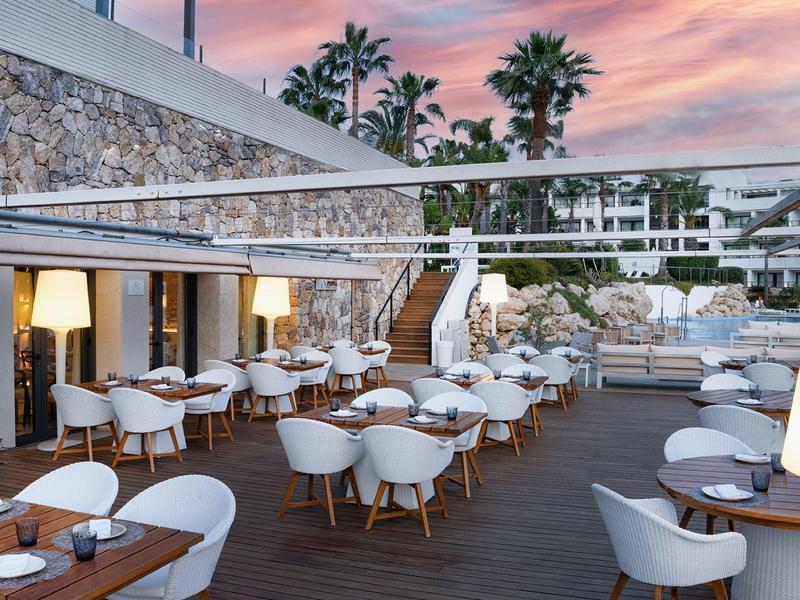 Terrasse d'hôtel avec chaises blanches et tables en bois au coucher du soleil avec des palmiers en arrière-plan.