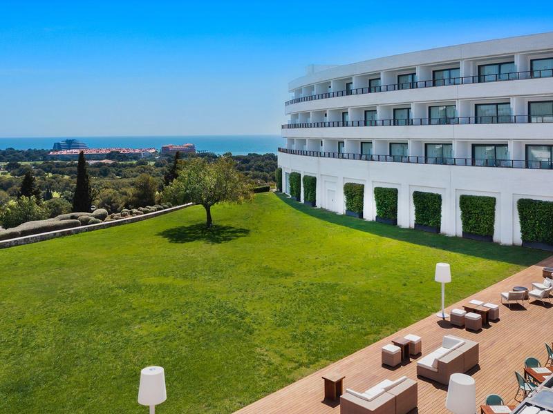 Bâtiment d'hôtel moderne avec chambres balcon, pelouse verte et vue sur la mer sous ciel dégagé.