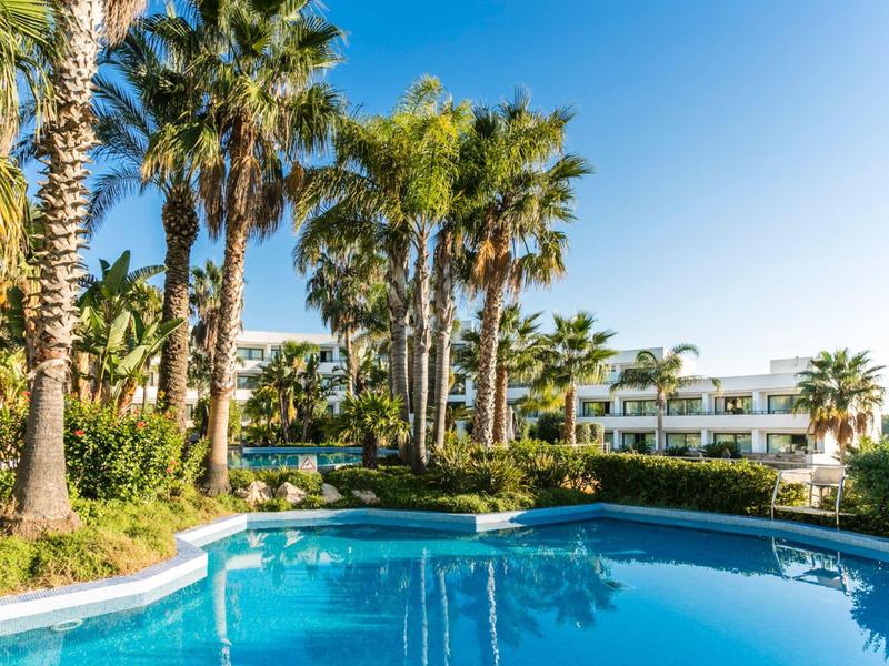 Une piscine bleue claire entourée de palmiers et de bâtiments d'hôtel sous un ciel bleu éclatant.