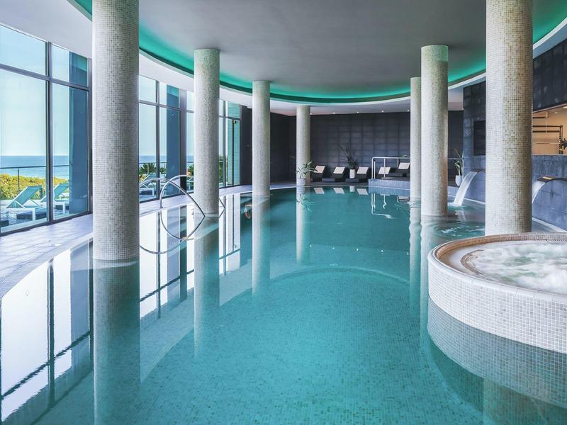 Piscine intérieure avec grandes fenêtres, piliers ronds et jacuzzi dans un hôtel moderne.