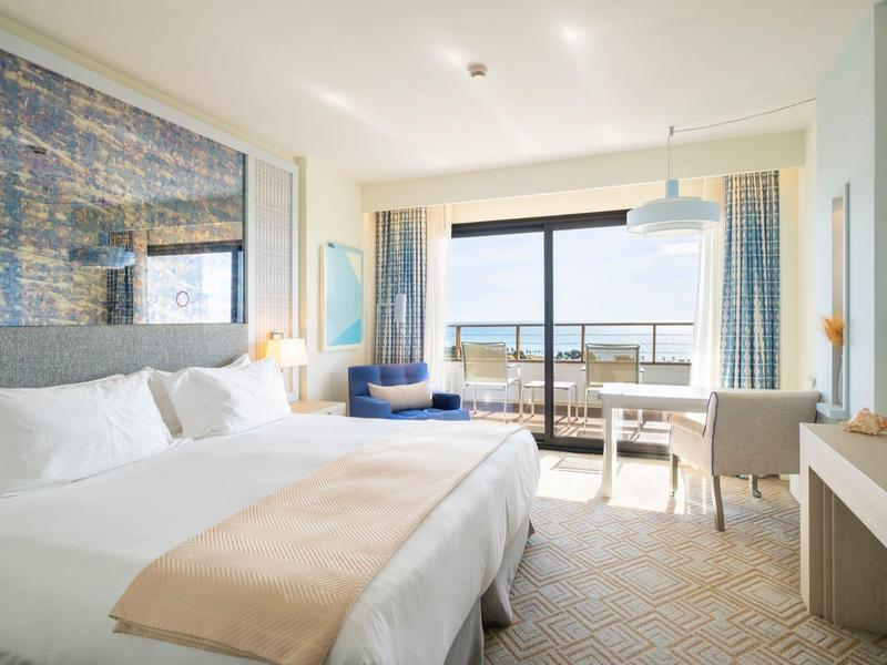 Chambre d'hôtel lumineuse avec grand lit, bureau et vue sur la ville par porte coulissante en verre.