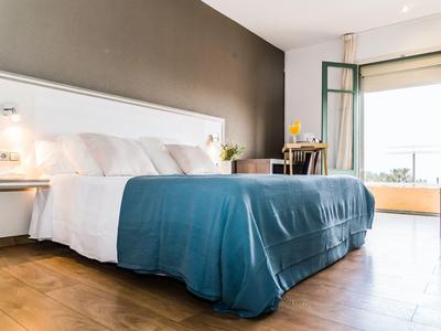 Bright hotel room with large bed, blue bedspread, wooden floor, and balcony with ocean view.