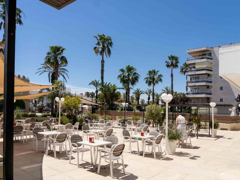 Sunny hotel terrace with tables, chairs, and palm trees in the background.