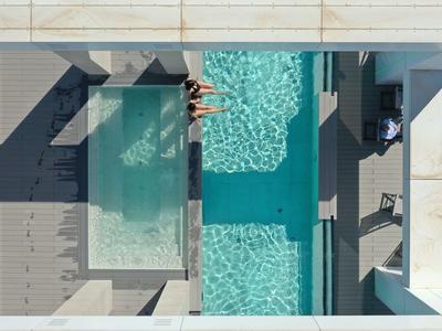 Tres personas nadan en una piscina exterior rectangular y moderna con tumbonas y sombrillas alrededor.
