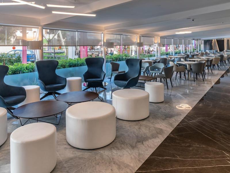 Moderna recepción de hotel con asientos, taburetes blancos redondos y grandes ventanas con vista al verde.