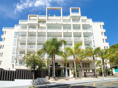 Bílá vícepodlažní hotelová budova s balkony a palmami vpředu.