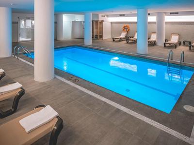 Moderní krytý bazén s modrým osvětlením, obklopený lehátky s bílými ručníky.