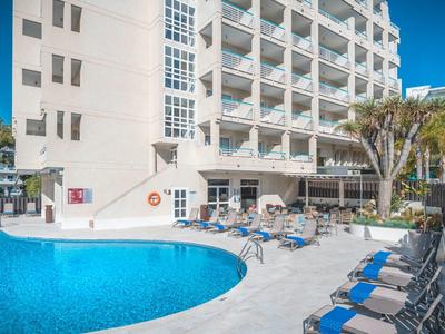 Hotelový bazén s modrými lehátky a slunečníky během slunečného dne.