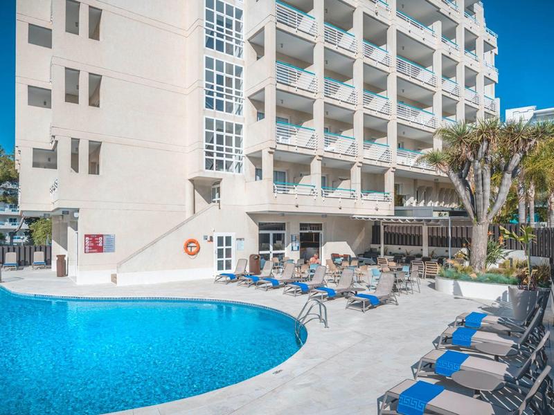 Hotelový bazén s modrými lehátky a slunečníky během slunečného dne.