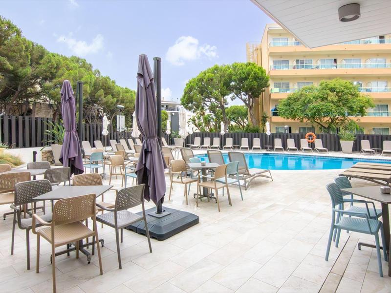 Hotelová bazénová zóna s židlemi, stoly a zavřenými slunečníky venku.