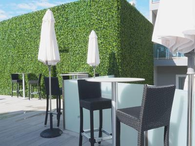 Zona esterna moderna con tavolo, sedie e ombrelloni bianchi davanti a siepe verde.