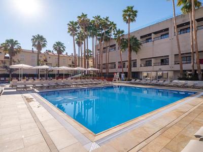 Piscina all'aperto con lettini e palme davanti a un edificio dell'hotel in una giornata di sole.