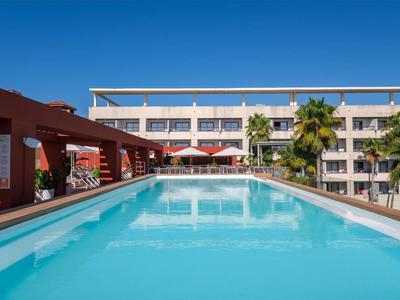 Piscina esterna moderna con acqua azzurra e ombrelloni davanti a un edificio alberghiero sotto un cielo limpido.