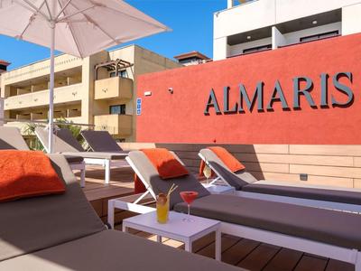 Lettini con cuscini arancioni sotto un ombrellone su una terrazza di fronte a un edificio rosso con la scritta ALMARIS