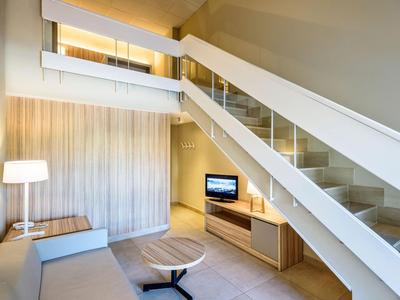Chambre d'hôtel moderne avec escalier, canapé, TV et intérieur lumineux aux tons neutres.