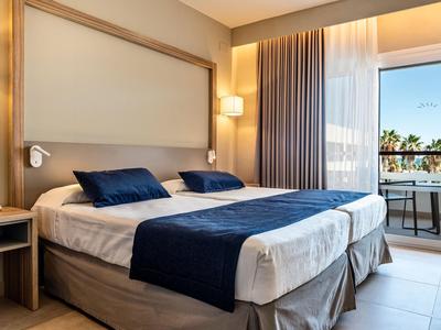 Chambre d'hôtel moderne avec grand lit double, literie bleue et blanche et balcon avec vue sur la mer.