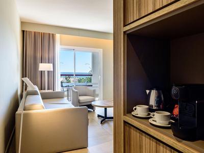 Coin salon moderne dans une chambre d'hôtel avec canapé, fauteuil et équipement café sur étagère en bois.
