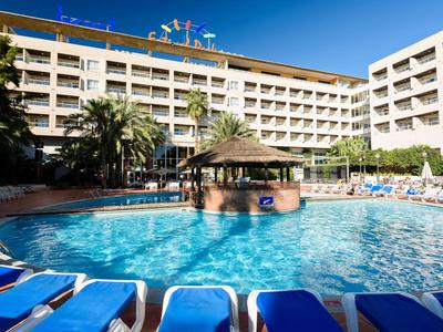 Ein Hotel mit angeschlossenem Pool, Sonnenliegen und Palmen bei sonnigem Himmel.