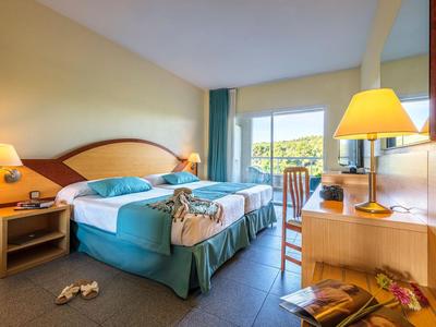 Jasny pokój hotelowy z podwójnym łóżkiem, szafkami nocnymi, biurkiem i tarasem z widokiem na zieleń.