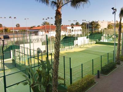 Ein sonniger Tennisplatz umgeben von grünen Zäunen, Palmen und Gebäuden im Hintergrund.