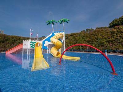 Buntes Kinderplanschbecken mit Rutschen, Wasserfontänen und Palmen-Dekor vor grünem Hügel.