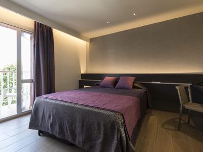 Modernes Hotelzimmer mit großem Bett, lila Bettdecke, Stuhl und Balkon mit Tageslicht.