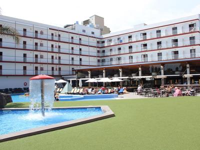 Hotel mit Außenpool, Wasserfontäne und Liegestühlen auf grünem Rasen bei Tag.