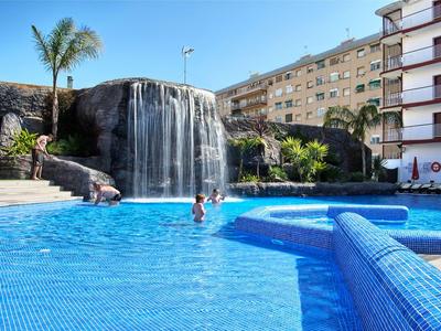 Hotelpool mit Wasserfall, umgeben von Felsen, Palmen und mehrstöckigen Gebäuden bei sonnigem Wetter.