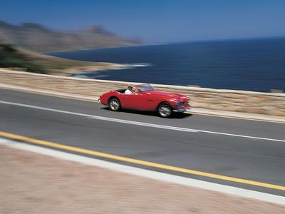 Roter Oldtimer fährt auf Küstenstraße mit Meer im Hintergrund.