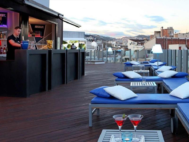 Dachterrasse mit blauen Liegen, weißen Kissen, Barbereich und Blick auf Stadt bei Abenddämmerung.