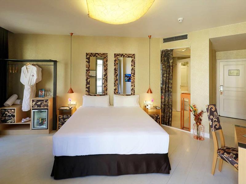 Helles Schlafzimmer mit großem Bett, zwei Spiegeln, Schreibtisch, Sesseln und einem Bademantel an der Wand.