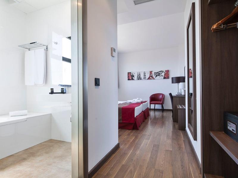 Moderní hotelový pokoj s dřevěnou podlahou, červeným křeslem a bílou koupelnou s vanou.