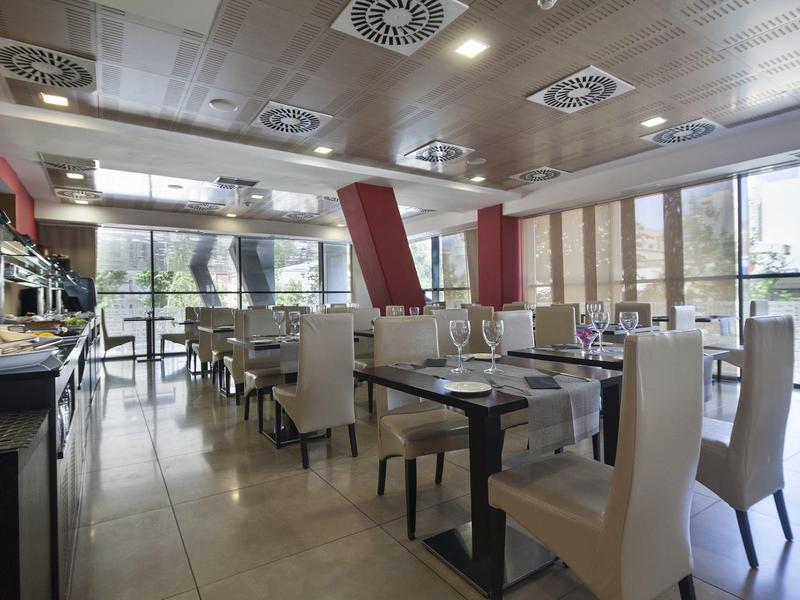 Moderní restaurace s bílými židlemi, dřevěnými stoly, velkými okny a stropním osvětlením.