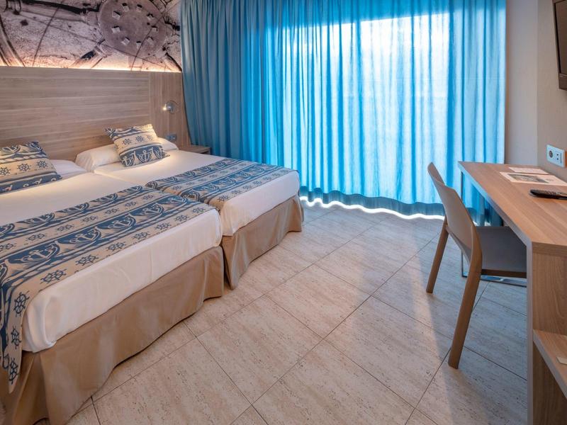 Chambre d'hôtel lumineuse avec deux lits simples et grande fenêtre avec rideaux bleus.