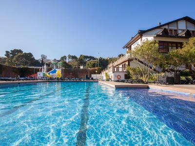Großer blauer Pool mit Wasserrutsche, umgeben von Bäumen und Gebäuden unter klarem Himmel.