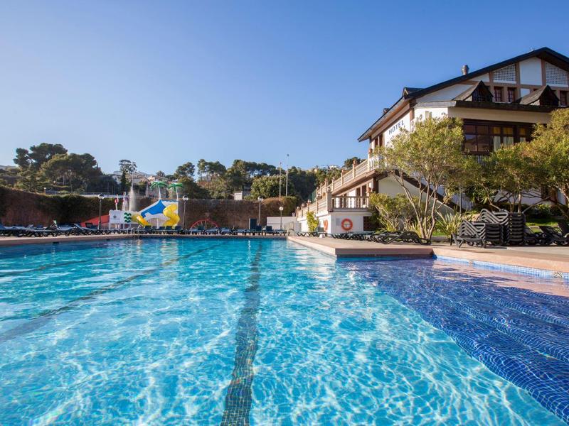 Großer blauer Pool mit Wasserrutsche, umgeben von Bäumen und Gebäuden unter klarem Himmel.