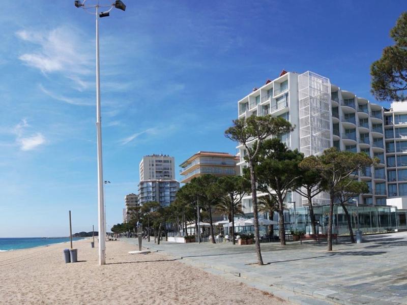 Sandy beach with promenade, trees, and several modern hotels by the sea under a blue sky