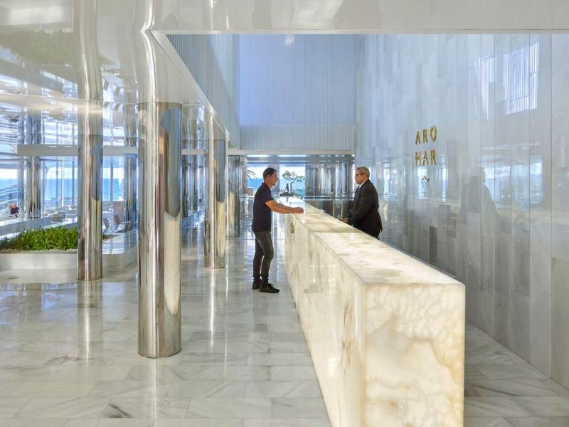 Brightly lit hotel lobby with marble reception desk and two people conversing.