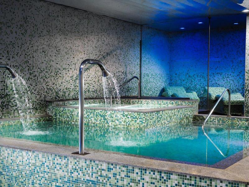 Binnenzwembad met mozaïektegels en blauw licht, met waterstralen en trappen.