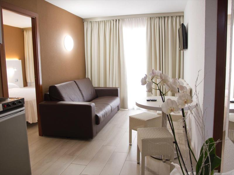 Nowoczesny pokój hotelowy z aneksem kuchennym, sofą, stołem jadalnym i sypialnią w tle