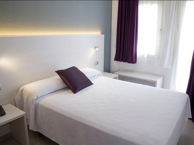 Jasny pokój hotelowy z białym łóżkiem, fioletowymi zasłonami i poduszką oraz stolikiem nocnym.