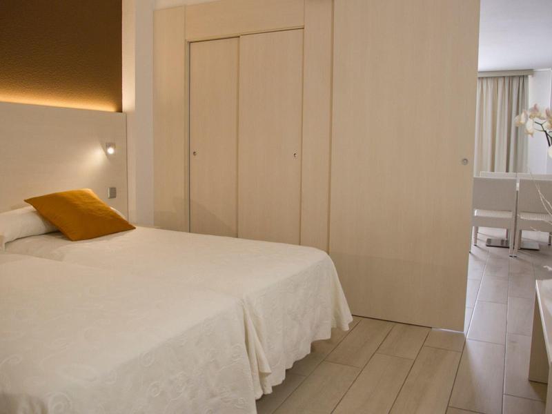 Minimalistyczna sypialnia z białą pościelą, pomarańczowymi poduszkami i jasnymi meblami drewnianymi.