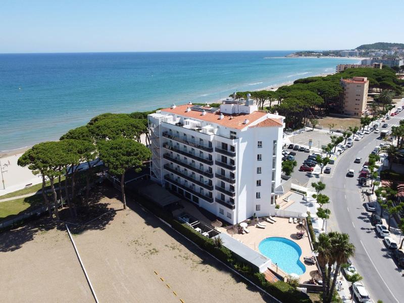 Edificio de hotel con piscina junto a la playa de arena y carretera, rodeado de árboles y mar al fondo.