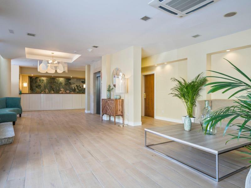 Recepción de hotel moderna y luminosa con mostrador, plantas verdes y áreas de descanso.